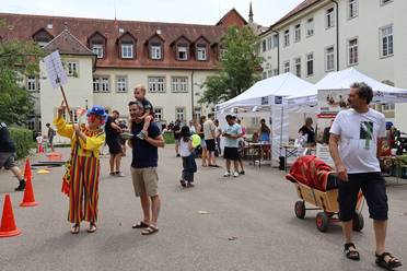 Jahresfest 2025 – Personen und Informationsstände im Schlosshof, eine bunt gekleidete Person zeigt auf ihr Schild, das sie an einem Stock trägt.