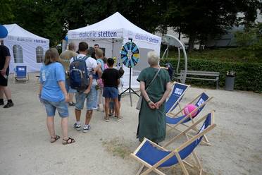 Jahresfest 2025 – Mehrere Personen stehen auf Sand vor einem Informationsstand im Park, neben ihnen stehen blaue Liegestühle, die zum Platz nehmen einladen.