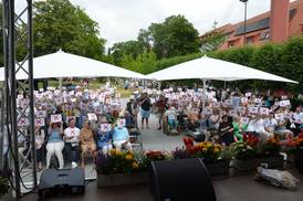 Jahresfest 2026 – Vor der Bühne sitzen zahlreiche Personen und halten je ein weißes Schild mit rotem Kreuz in den Händen Jahresfest 2026 – Vor der Bühne sitzen zahlreiche Personen und halten je ein weißes Schild mit rotem Kreuz in den Händen