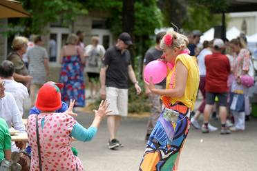 Jahresfest 2025 – Zwei Damen im bunten Kleidungsstücken unterhalten die Besucher*innen, eine von ihnen bläst einen Luftballon auf.